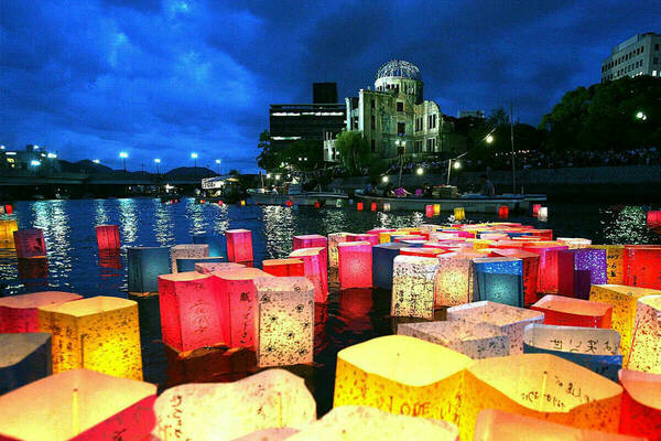 At dusk, colorful floating paper lanterns with messages written on them illuminate the waters in front of the Hiroshima Peace Memorial. In the background, the skeletal dome of the memorial is visible, lit from below. A bridge and other buildings are also in the background.