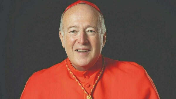 Cardinal Timothy Dolan, Archbishop of New York, smiles against a dark background. He wears a red zucchetto and cassock.