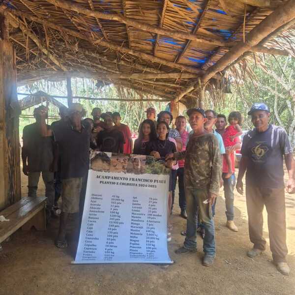 Diverse group under a thatched roof holding a banner for Acampamento Francisco Piauí's 2024-2025 crop plan.