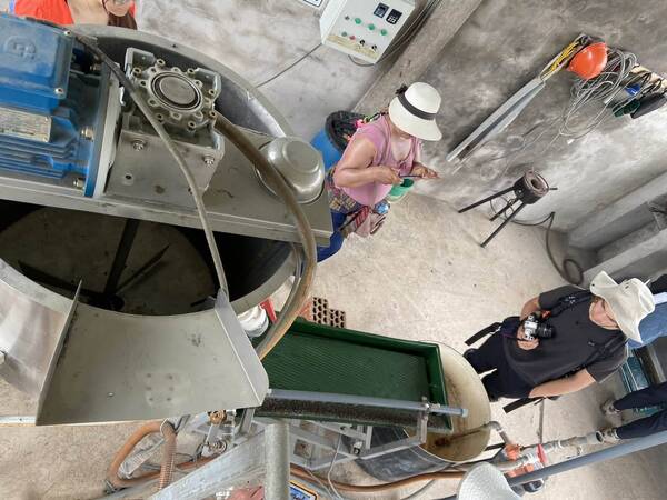 Woman in white hat, pink shirt uses phone near large industrial machine with blue motor and green tray. Man with camera watches.