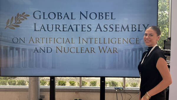 Smiling woman in black top next to screen: Global Nobel Laureates Assembly on Artificial Intelligence and Nuclear War.